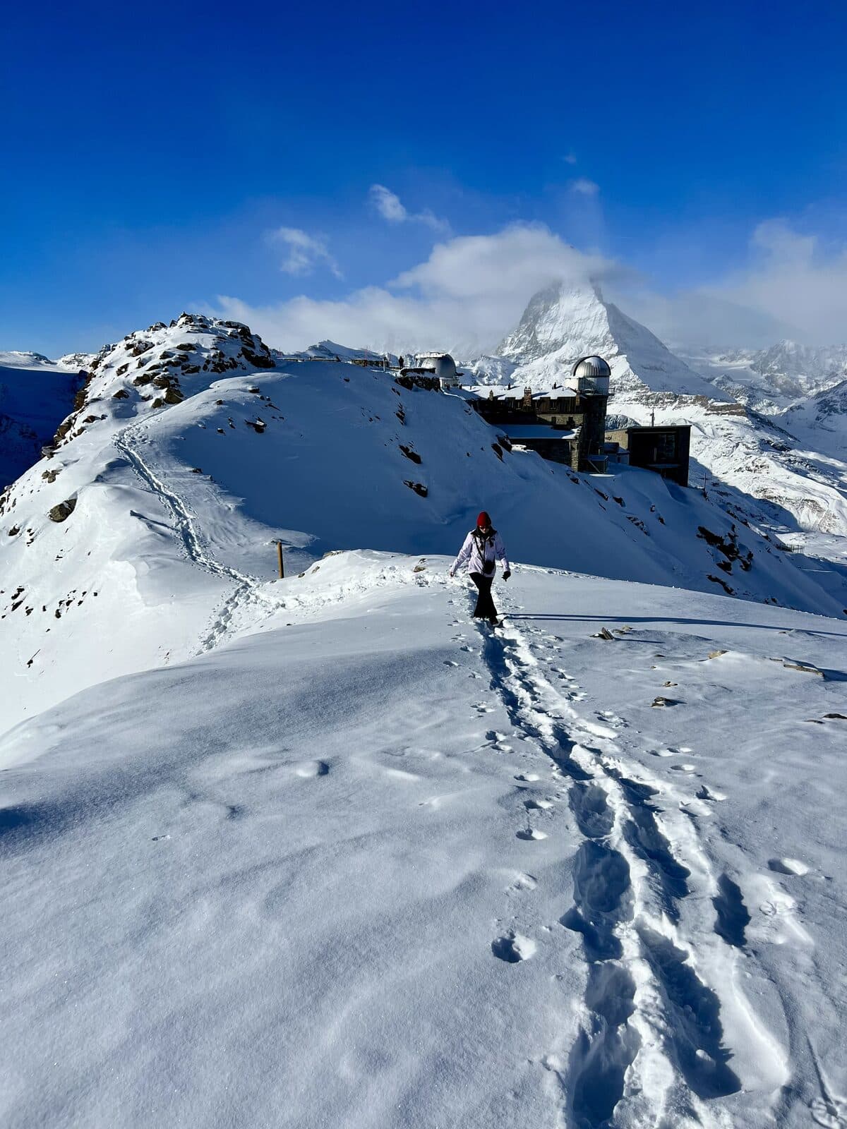 Dylan's wife hiking along a snowy ridge at Gornergrat, with the Matterhorn and the observatory domes behind